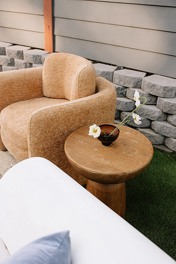 Wedding lounge seating with an upholstered armchair beside a round wooden side table topped with a bud vase of white flowers on grass by a stone wall
