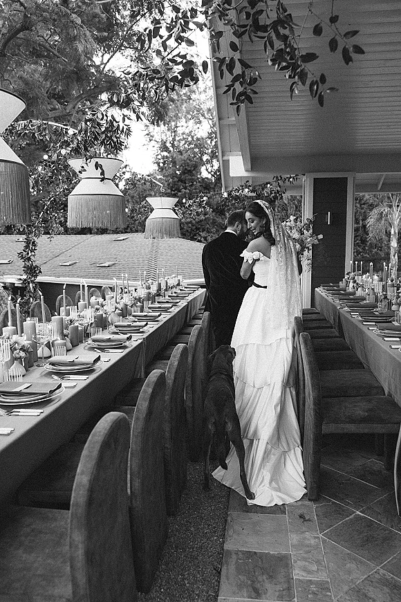 Newlywed couple portrait from behind, bride looking back in long veil and gown beside a dog at candlelit outdoor patio banquet tables﻿