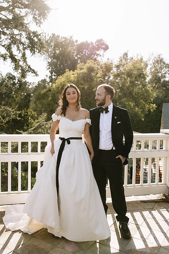 Couple portrait of bride and groom holding hands on a sunlit balcony, bride in off-shoulder gown with black sash, groom in tuxedo