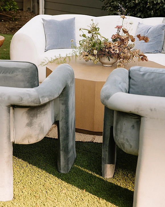 Lounge seating area with wedding lounge furniture featuring a white sofa, blue velvet armchairs, round rug, and low floral centerpiece on a lawn