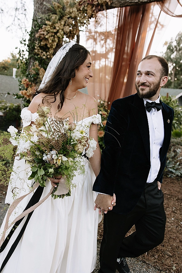 Couple portrait of bride and groom holding hands, bride looking at groom, veil and bouquet, walking a garden path by trees and roofline