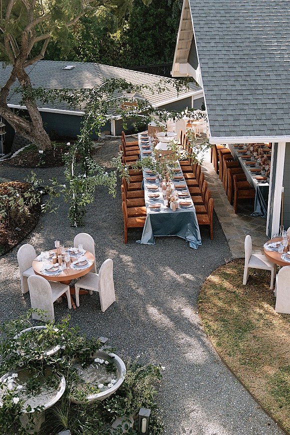Outdoor reception tablescape with a long banquet table setup, taper candles and greenery garland beneath string lights in a courtyard