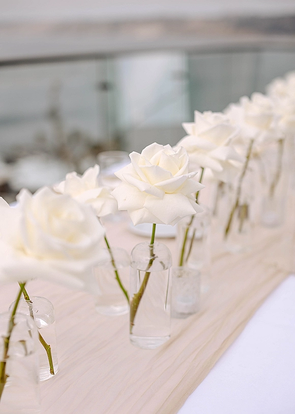 Wedding centerpiece with white rose centerpiece in clear glass bud vases and water-filled cylinders on a light wood runner, indoor venue backdrop