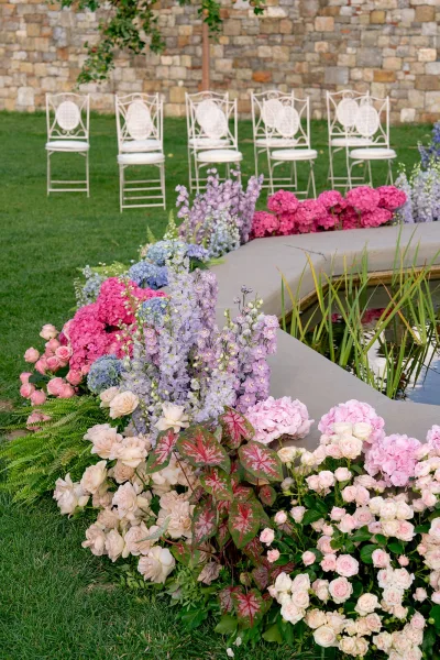 Ceremony floral decor with outdoor ceremony flowers lining white chairs, hydrangeas and roses edging a pond beside a stone wall backdrop