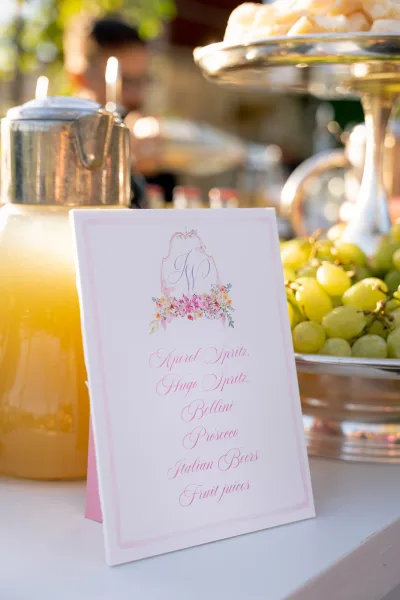 Wedding drink station with a wedding drink menu sign, glass dispenser and silver container, grapes and citrus, set against outdoor greenery and blurred guests