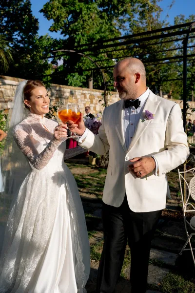 Wedding toast as bride in lace long-sleeve dress and groom in white dinner jacket clink orange cocktails in sunny garden by stone wall