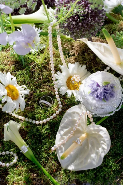 Wedding rings flatlay with diamond rings, pearl necklace, and beaded ring box nestled on green moss with calla lilies, daisies, and purple allium