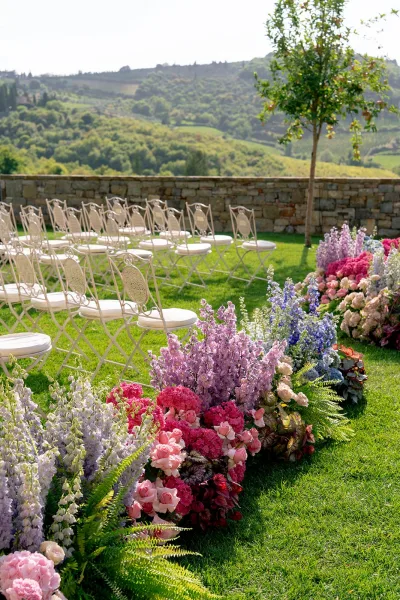 Ceremony aisle decor with outdoor ceremony setup, pink and purple florals and ferns lining chairs on a lawn with rolling hills beyond