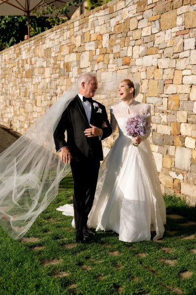Wedding bride portrait with a high-neck lace dress and long veil, holding a purple bouquet beside a black tuxedo by a stone wall outdoors