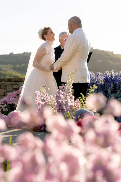 Wedding vows during an outdoor wedding ceremony as bride in lace sleeves and veil holds hands with groom in white jacket on hillside terrace