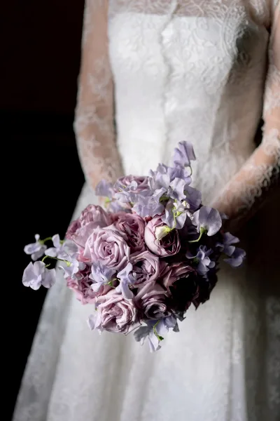 Bridal bouquet with lavender rose bouquet and sweet pea flowers held against a bridal gown with lace sleeves on a dark interior backdrop