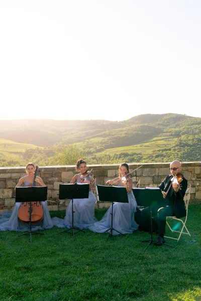 Wedding ceremony musicians in a string quartet wedding perform with sheet music on stands, seated on a sunny lawn by a stone wall