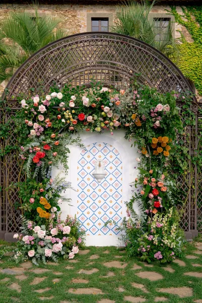 Wedding ceremony backdrop with a floral ceremony arch of roses and greenery on a trellis beside a tile fountain in a stone courtyard
