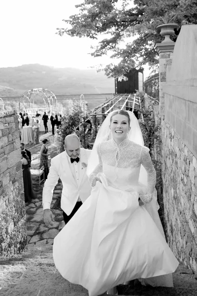Bride portrait in black and white wedding portrait style, lifting her lace long-sleeve gown on stone steps with groom behind on terrace