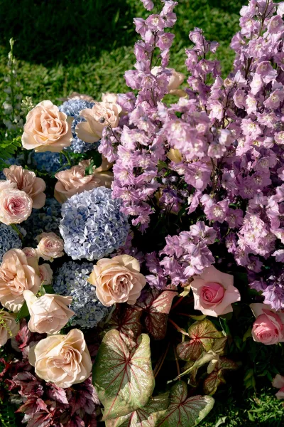 Wedding florals in a garden wedding flowers arrangement with roses, hydrangea, purple delphinium, and greenery on a grass lawn