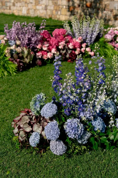Wedding ceremony florals with ground floral arrangements of hydrangeas, delphinium, roses, and greenery on a lawn by a stone wall