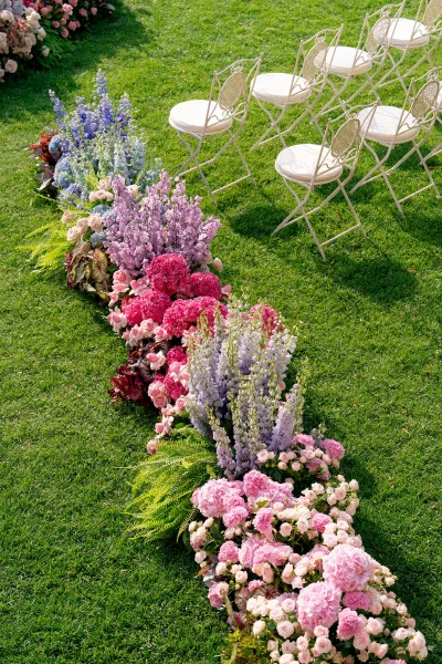 Ceremony aisle flowers lining a grass lawn aisle with roses, hydrangeas, ferns, and white metal chairs with cushions beside them