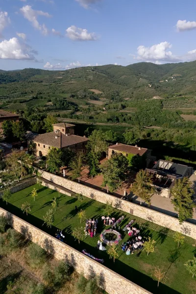 Outdoor ceremony setup with circular wedding ceremony seating, floral arch, white aisle runner, parasols, on a stone terrace lawn with hills beyond