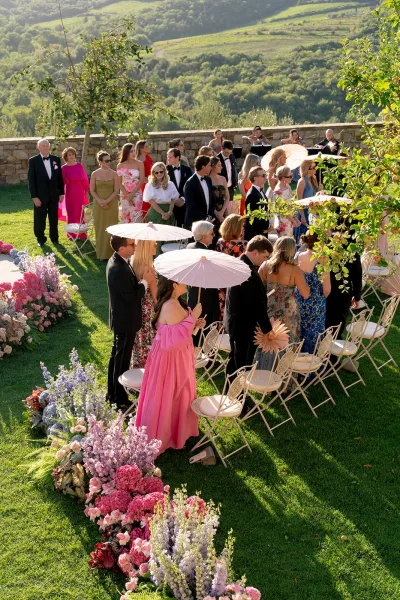 Ceremony guests in formal attire seated on white folding chairs, some with white parasols, listening to a string quartet on a garden lawn with stone wall backdrop