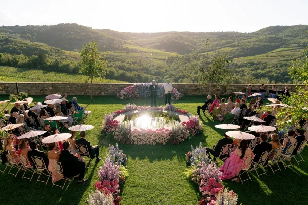 Ceremony setup with outdoor wedding ceremony seating around a circular reflecting pool, pastel florals and white parasols on a sunlit lawn with hills