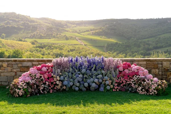 Wedding ceremony backdrop with floral ceremony backdrop of hydrangeas, roses, delphinium, and greenery before a stone wall and valley hills