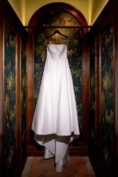 Wedding dress on hanger, strapless wedding dress with clean satin silhouette in an arched alcove with patterned wallpaper and uplighting