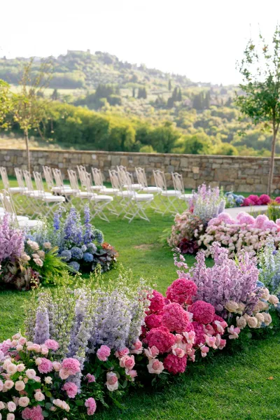 Ceremony aisle decor with floral lined wedding aisle of pink hydrangeas, roses, and delphinium beside white folding chairs on a lawn
