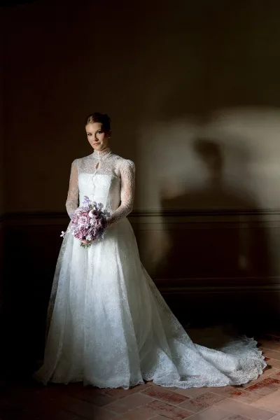 Bridal portrait in a long sleeve lace wedding dress, holding a lilac bouquet by moody indoor wall with soft window light shadows