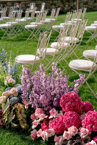 Ceremony aisle florals with outdoor ceremony aisle flowers in low clusters of roses, hydrangea, and greenery beside white chairs on lawn by stone wall