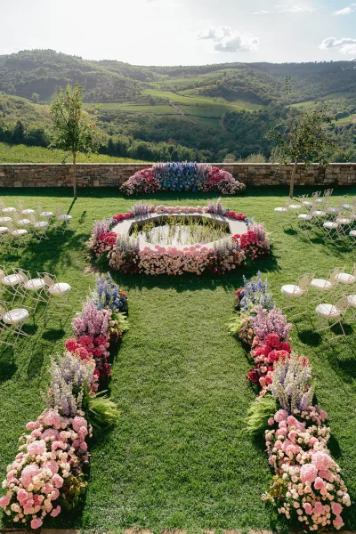 Ceremony setup for an outdoor wedding ceremony with floral aisle arrangements circling a reflecting pool with floating candles on a lawn by vineyards