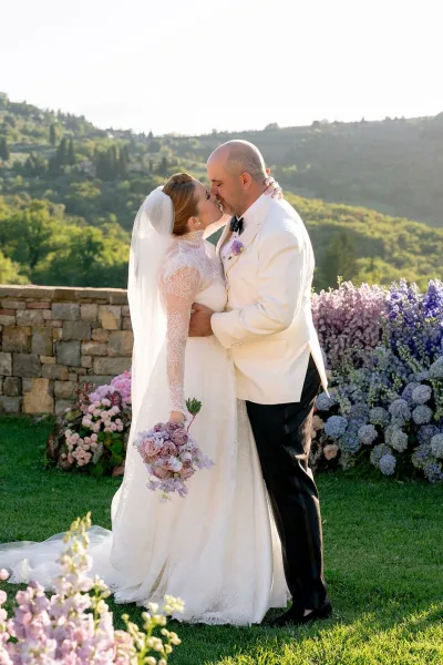 Wedding kiss portrait of bride and groom kissing, her veil trailing as he wears a white tuxedo jacket before a stone wall hillside backdrop