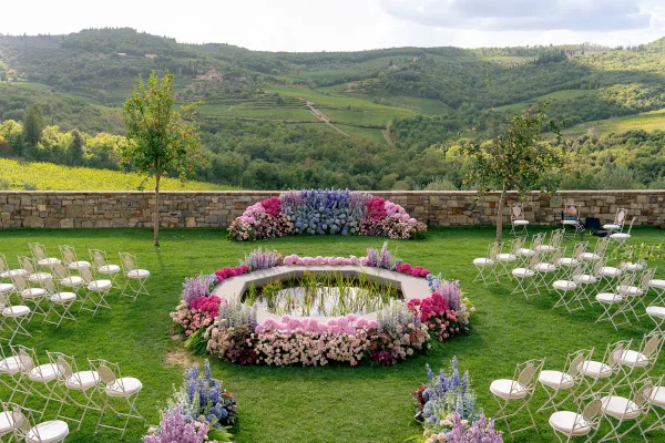 Ceremony setup for outdoor wedding ceremony with white folding chairs circling a reflecting pool, pastel florals, and vineyard hills beyond
