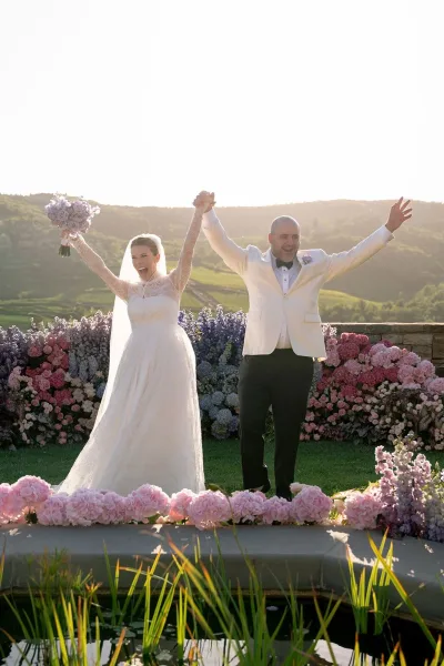 Just married couple in wedding recessional pose, arms raised as bride lifts bouquet and groom cheers at sunset by pond and hills