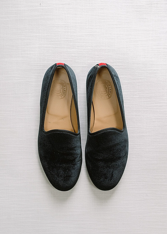Groom wedding shoes, black velvet loafers with red heel tabs resting on a white fabric surface in a clean wedding day detail flat lay