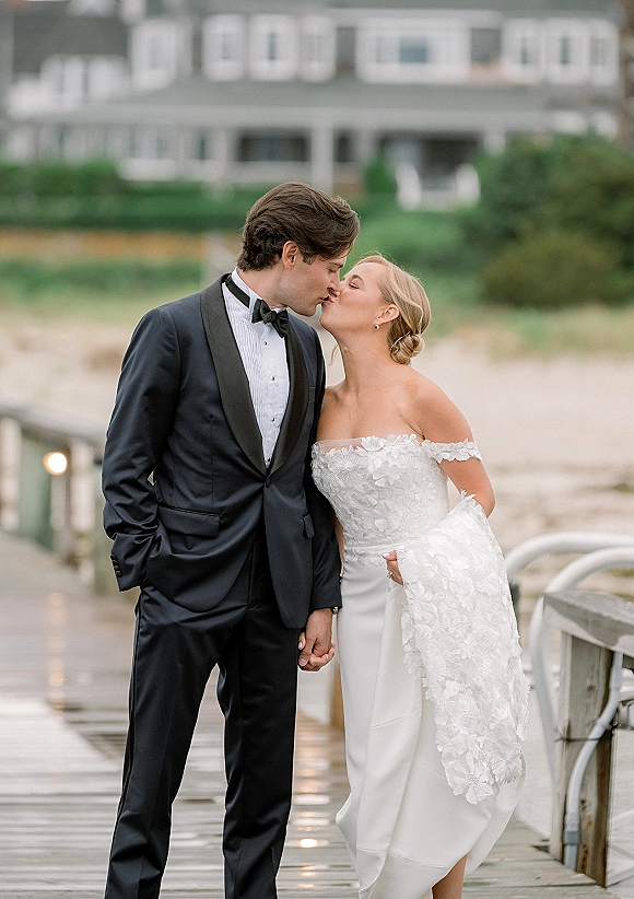 Wedding kiss portrait of bride and groom kissing on a wooden dock, her strapless lace dress and his black tuxedo by the water