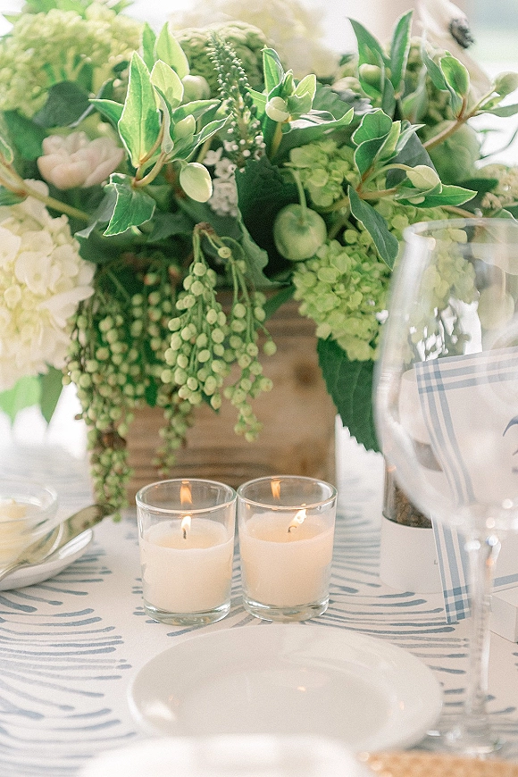 Reception tablescape with a wedding table centerpiece of green hydrangea and greenery in a wooden vase, with votive candles, plates, and wine glass in bright indoor light