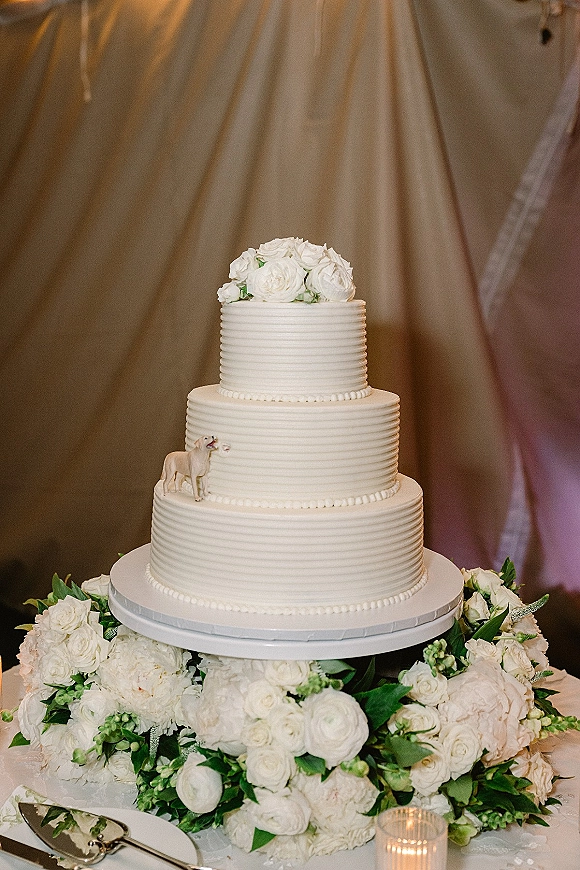 Wedding cake three tier wedding cake with white roses and greenery, dog topper on stand with knife, against draped fabric and string lights