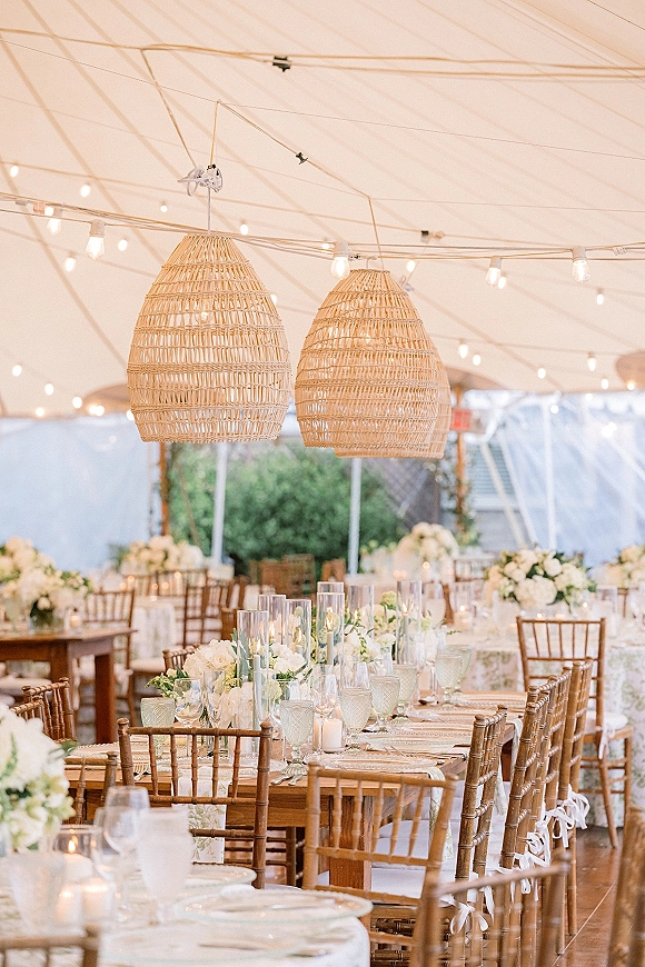 Reception tablescape on a farm table wedding reception with white florals, candle cylinders, glassware, and rattan pendant lights under a tent canopy