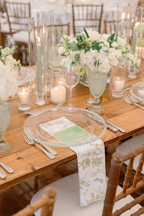 Reception tablescape on a wood farm table wedding setup with clear glass chargers, mint place cards, floral napkins, white centerpieces, and candles