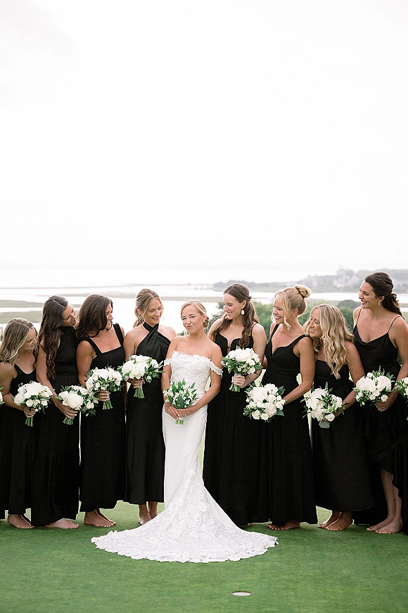 Bridesmaids portrait of bride with bridesmaids in black dresses holding white rose and greenery bouquets on a coastal lawn by the ocean horizon