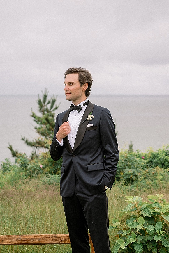 Groom portrait in a black tuxedo groom look, hand in pocket with boutonniere and bow tie by the ocean under a cloudy sky