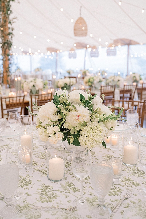 Reception tablescape with wedding table centerpiece of white blooms and greenery, candlelight in hurricane vases under string lights in a tent