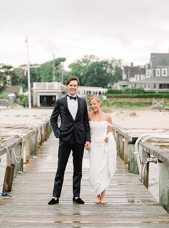Couple portrait of bride and groom holding hands on a wooden dock, barefoot bride in strapless lace dress beside tuxedoed groom by the water