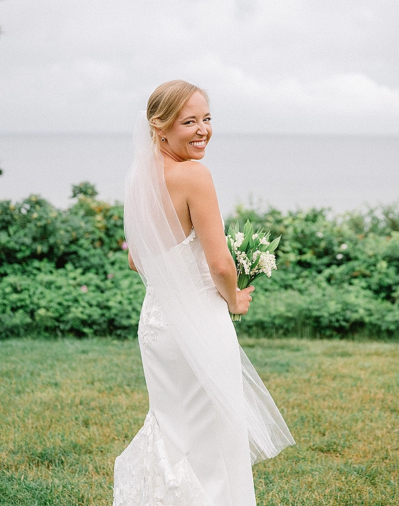 Bridal portrait of a bride looking back, smiling in a strapless lace gown with veil and lily of the valley bouquet by the ocean lawn
