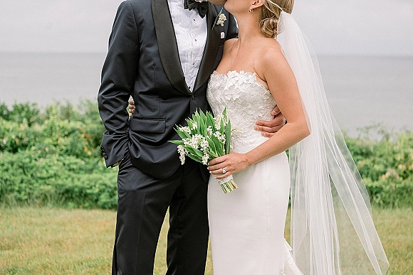 Wedding couple portrait of bride in strapless lace dress and veil kissing groom in black tuxedo, holding bouquet by calm waterfront lawn
