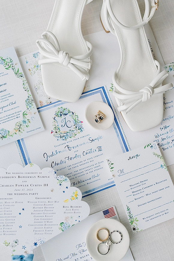 Invitation suite with blue floral wedding invitations, RSVP card, rings, earrings, ring box, and white bridal shoes on linen fabric surface