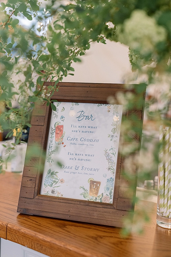 Wedding bar sign in a wood frame with printed cocktail menu and floral illustrations on a wooden counter, greenery blurred behind
