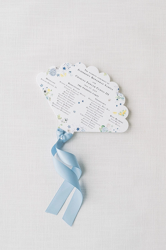 Wedding program with scalloped edge and floral illustration, tied with a blue ribbon bow and satin tail on white linen flat lay background