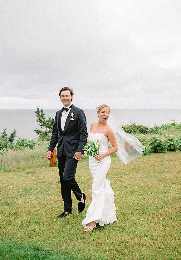 Couple portrait of bride and groom walking hand in hand, veil blowing as she holds a white bouquet on a coastal lawn by the ocean