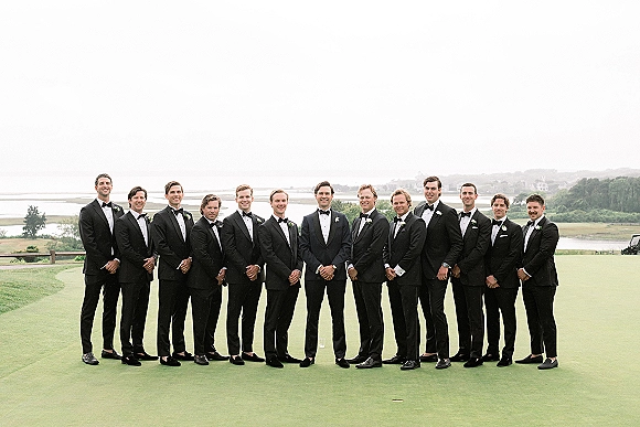 Groomsmen portrait of the groom with groomsmen in black tuxedos and bow ties on a golf course green by water under cloudy sky